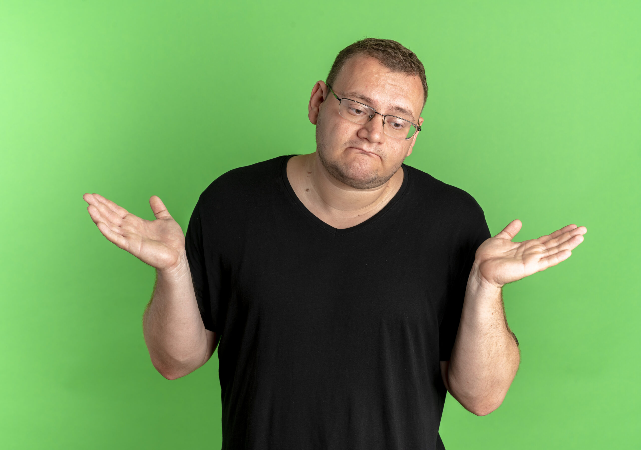 overweight man in glasses wearing black t-shirt looking confused and uncertain spreading arms to the sides having no answer standing over green background